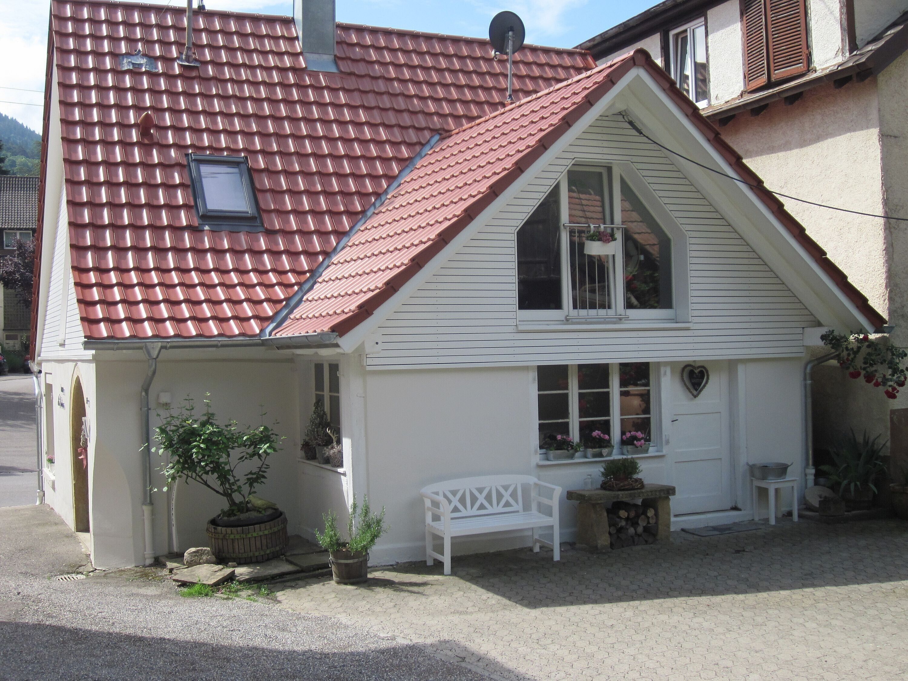 Une maison avec un toit recouvert de tuiles rouges, une façade blanche, un petit porche avec un banc et un décor en forme de cœur sur le mur.
