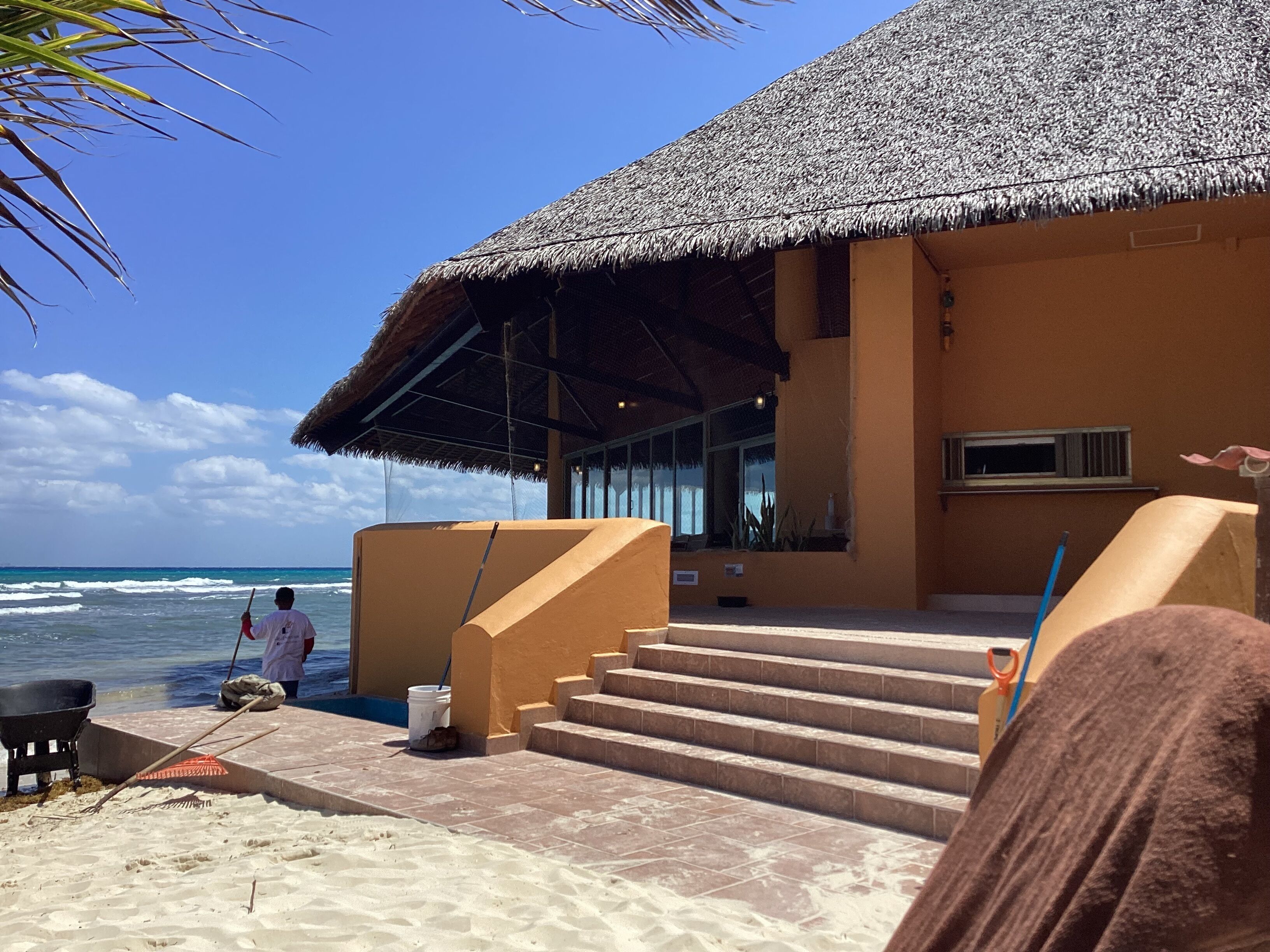 Un bâtiment situé au bord de la plage, doté d’un toit de chaume, d’un escalier menant à l’entrée et offrant une vue sur l’océan.