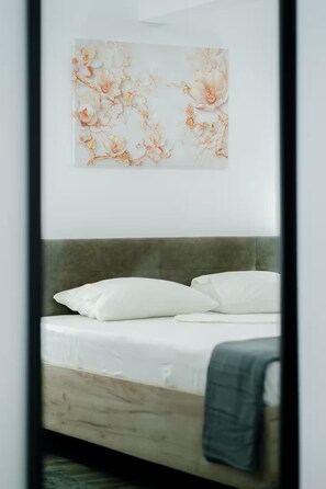 Cama com roupa de cama branca e cabeceira cinza em quarto com obra de arte na parede.