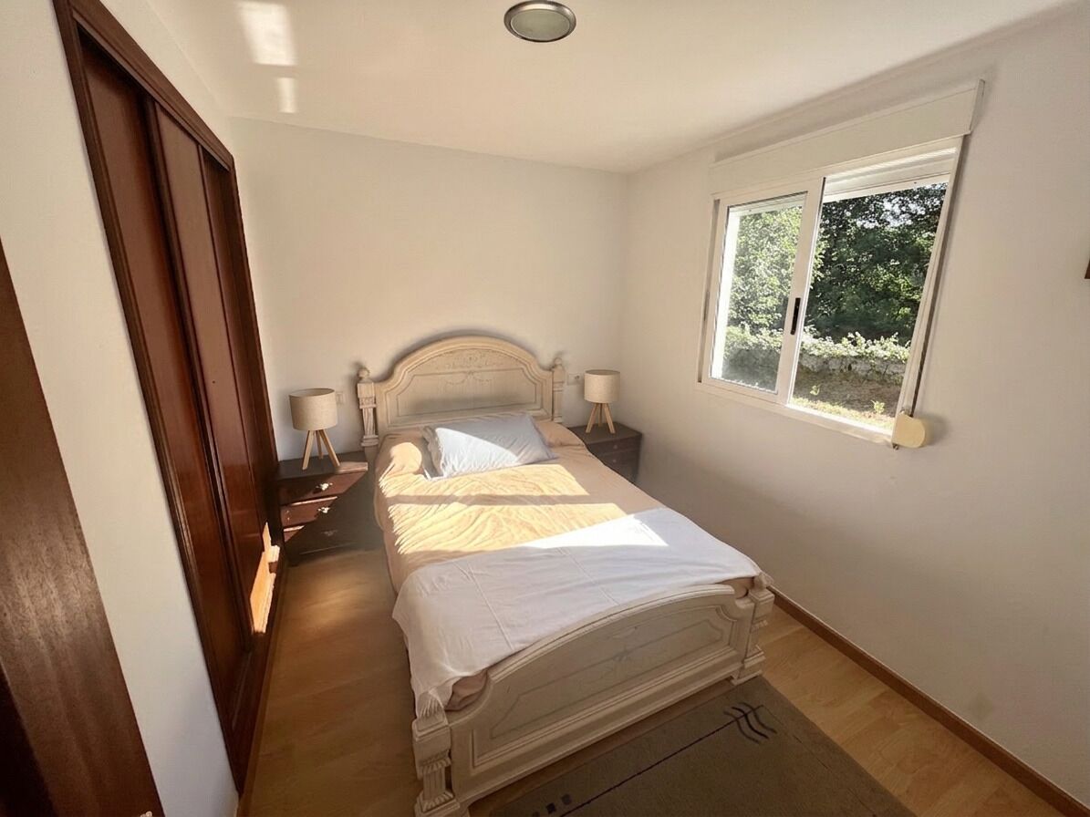 A bedroom with a single bed, bedside tables, a window with a view of greenery, and a door.