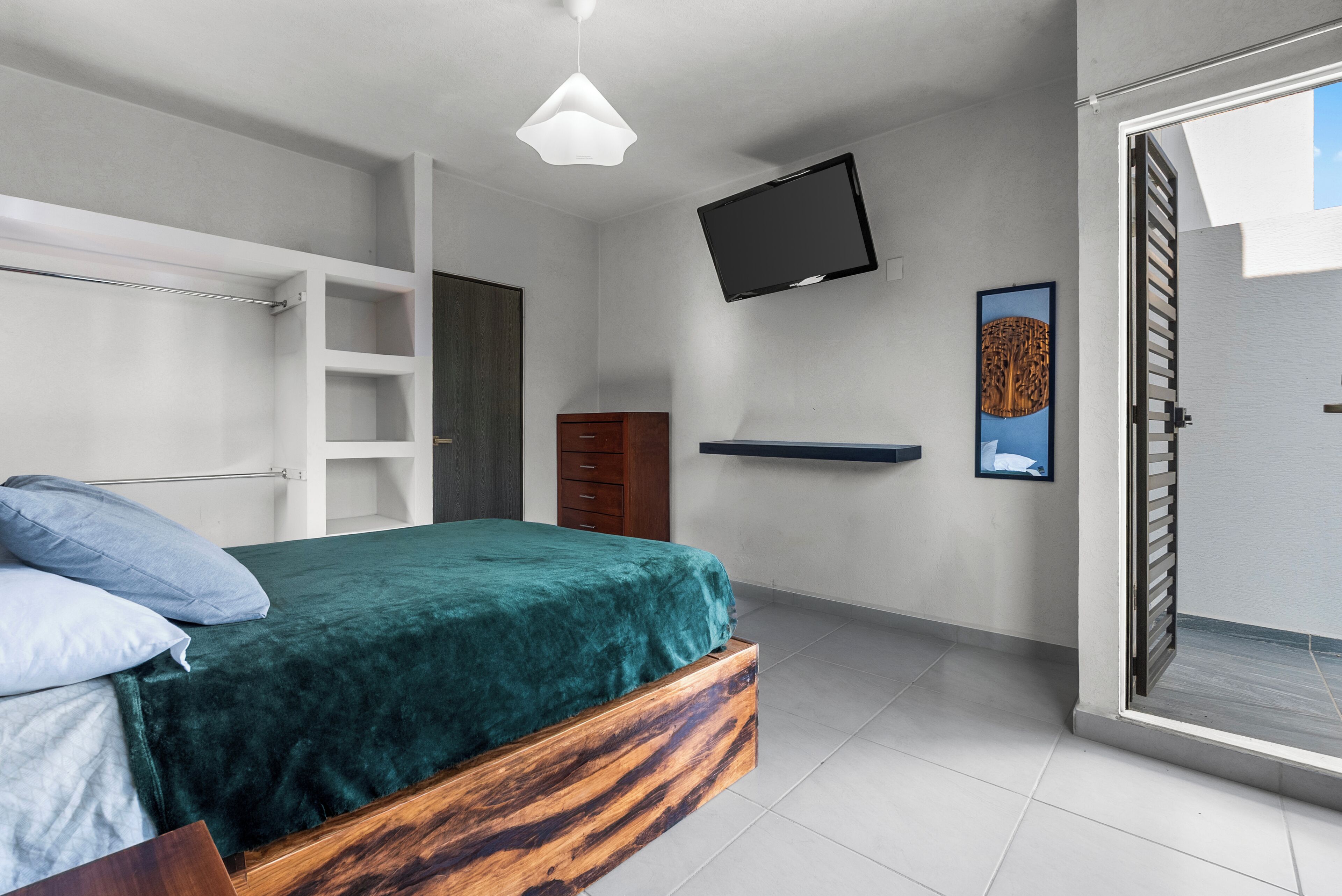 Un dormitorio con cama, televisor montado en la pared, armario de madera y puerta corrediza que da a un espacio exterior.