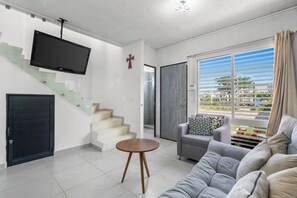 Un salon moderne avec un escalier, un téléviseur à écran plat, une décoration en forme de croix et une grande fenêtre donnant sur un paysage verdoyant.