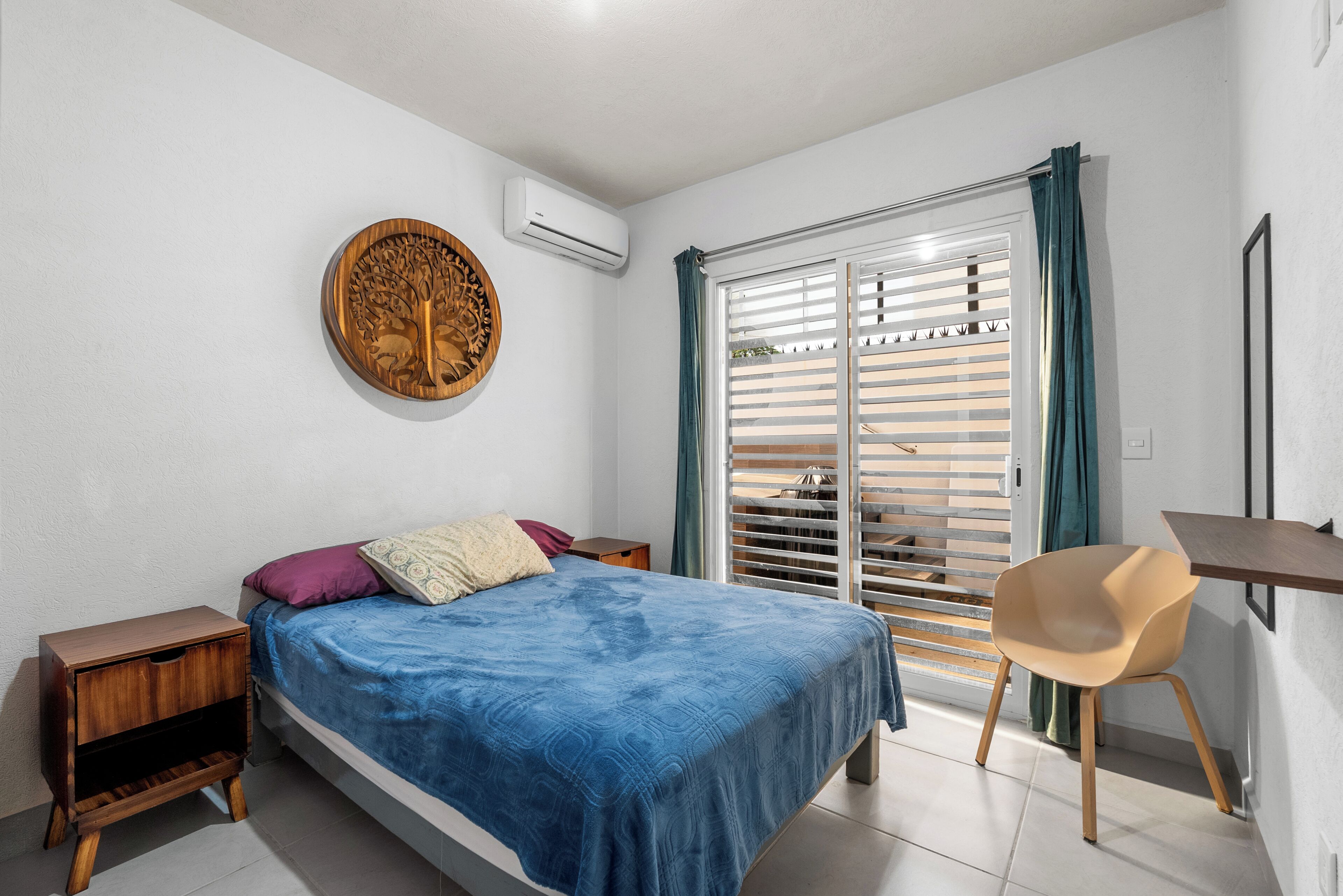 Un dormitorio con una cama, una mesita de noche de madera, un escritorio con una silla, un ventanal con persianas y un equipo de aire acondicionado.