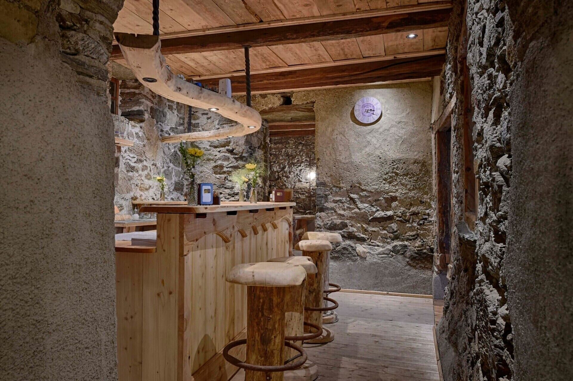 A rustic interior with a wooden bar and stools, stone walls, and a clock on the wall.