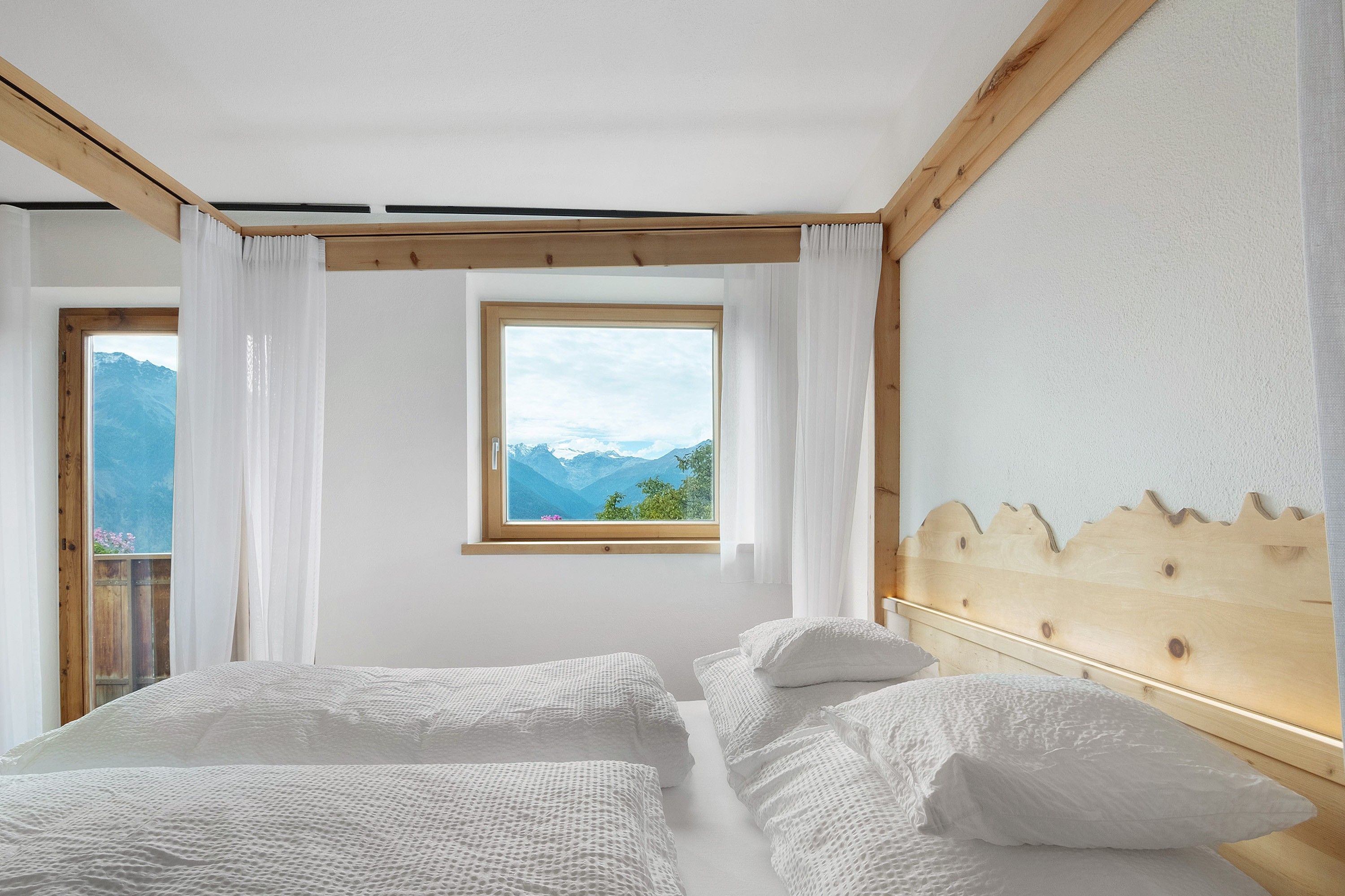 Un dormitorio con dos camas, ventana con vistas a la montaña, vigas de madera y cortinas blancas.