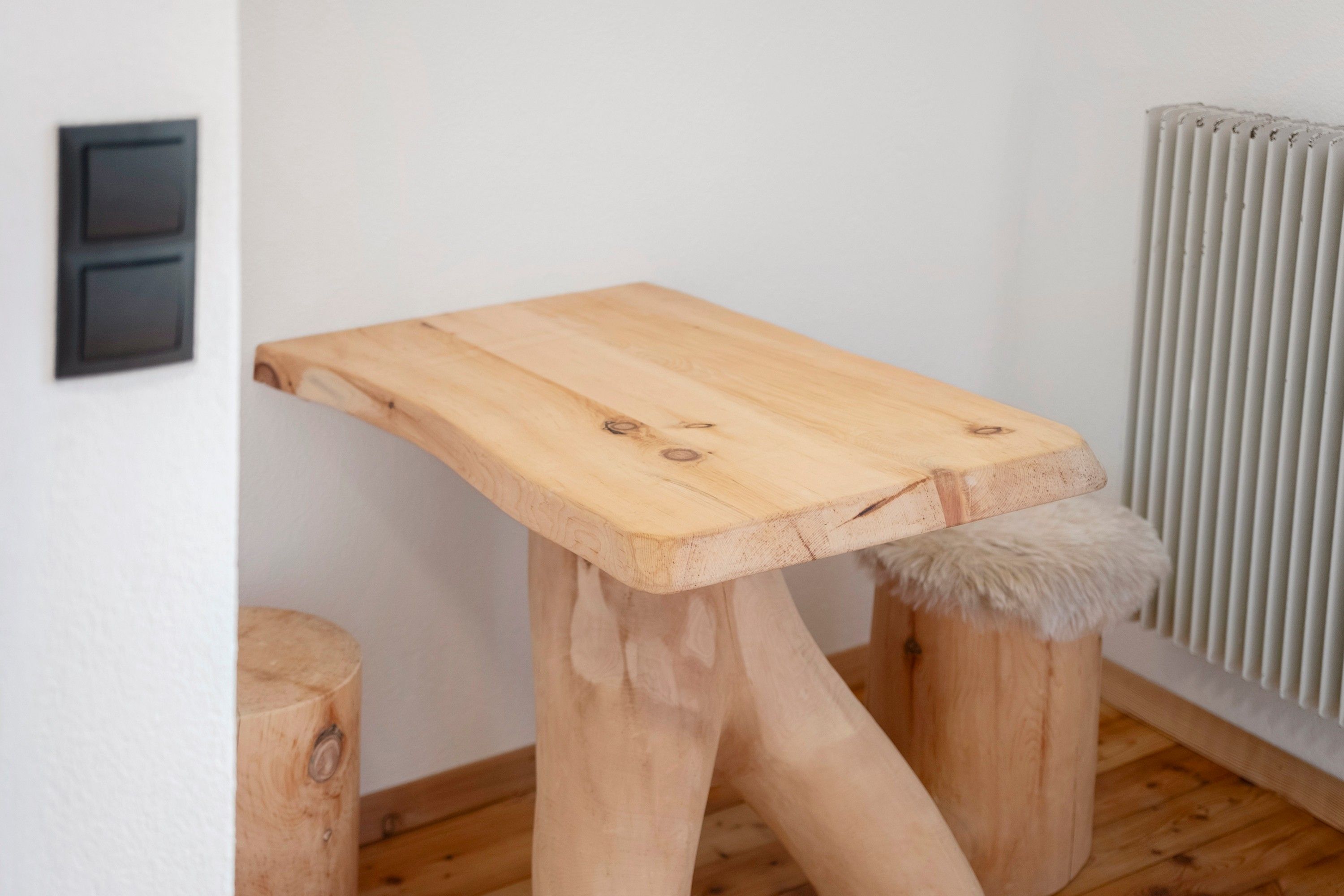Eine Katzenkralle aus Holz mit flacher Ablage und einem fellartigen Fußhocker, platziert vor einer Wand mit Heizkörper und Stromverteiler.