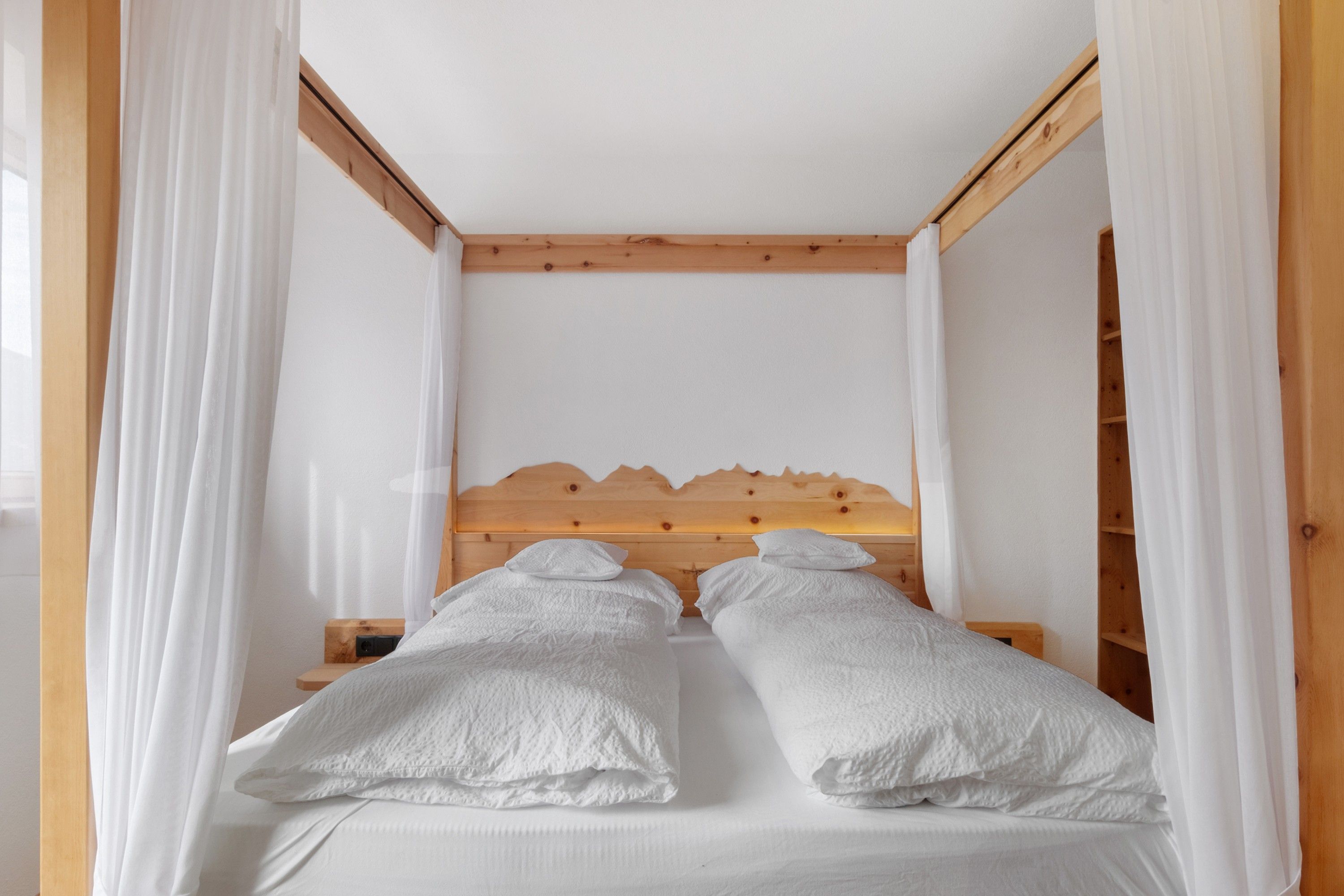 A bedroom with a wooden headboard, two white beds with pillows, and sheer white curtains.
