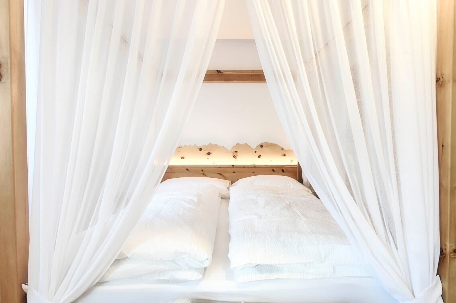 A bed with white linens and a wooden headboard, surrounded by sheer white curtains.