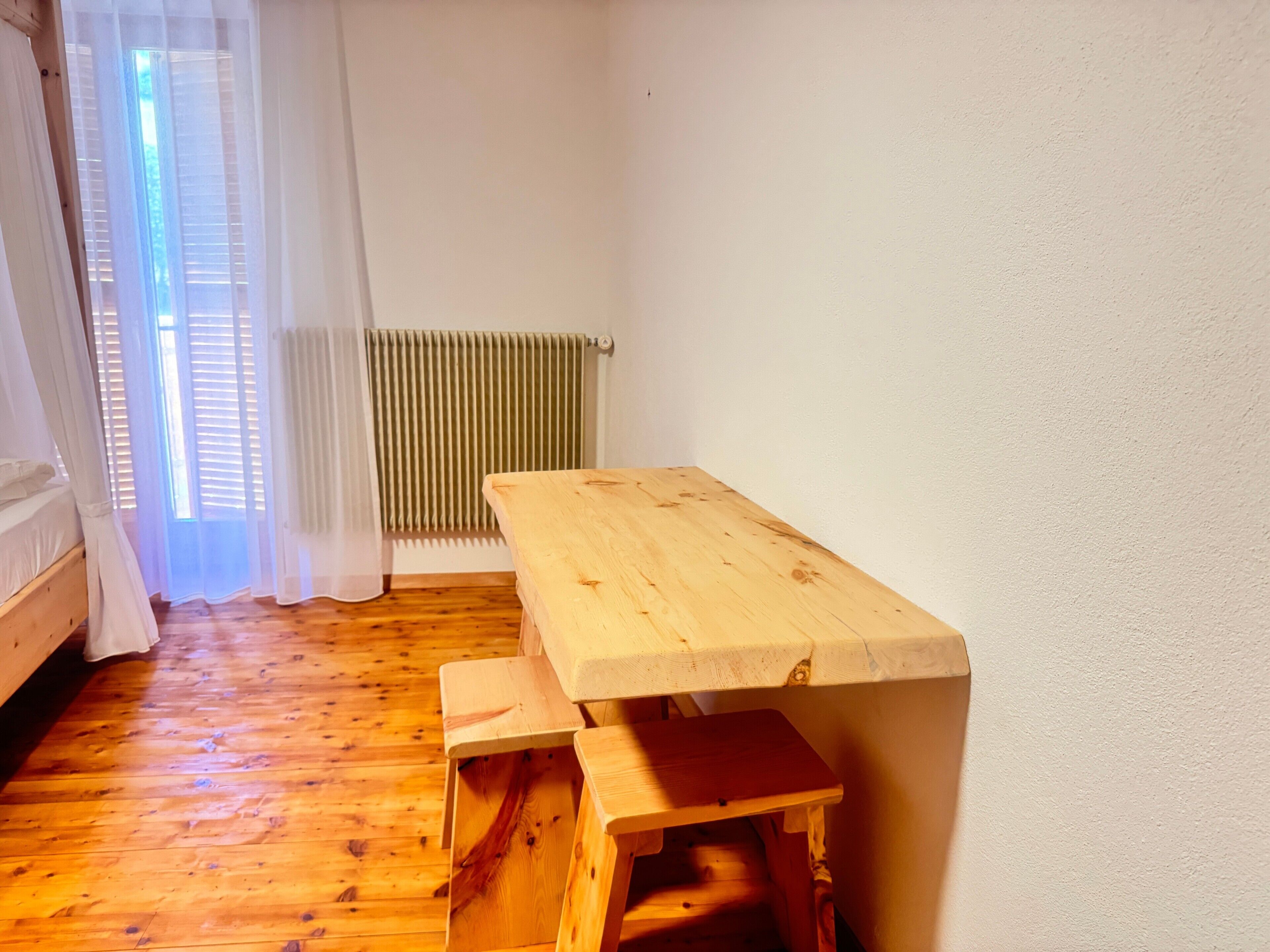 A room with a wooden floor, a wooden table with benches, a radiator, and a window with curtains.