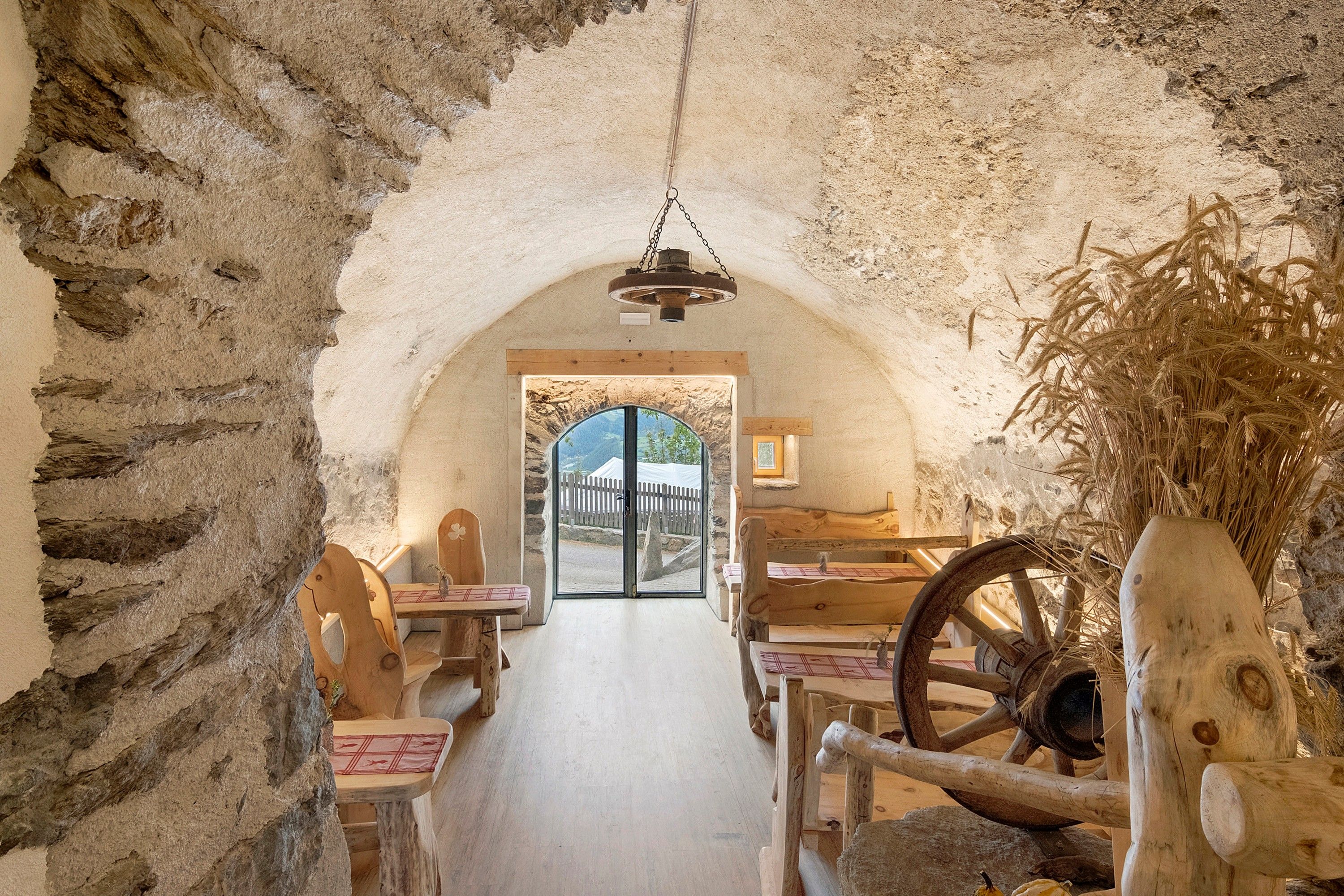 Un intérieur rustique avec des murs en pierre, des meubles en bois et une vue sur l’extérieur, visible à travers une porte en arc de cercle.