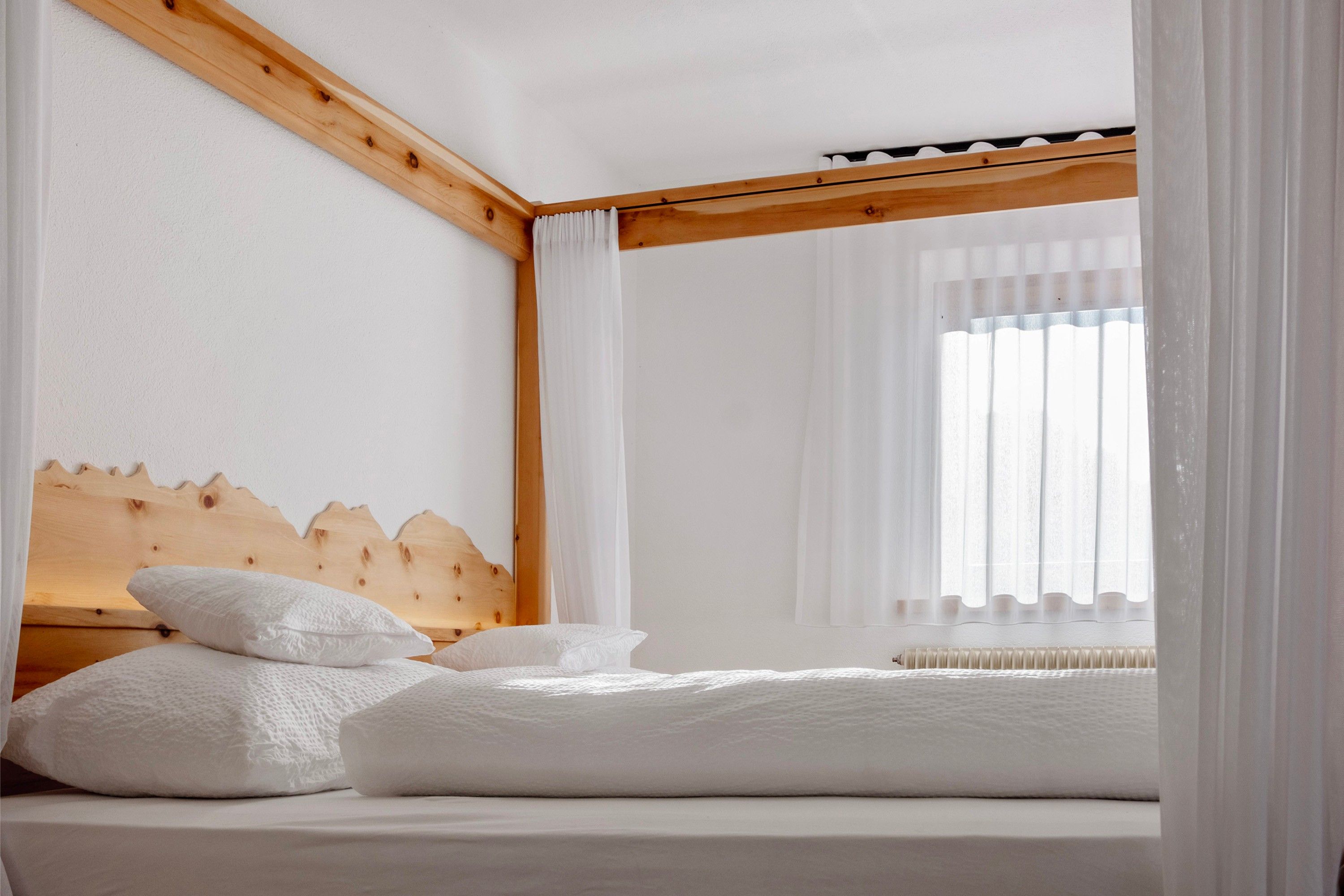 Ein Schlafzimmer mit einem Holzkopfende, weißer Bettwäsche und einem Fenster mit durchsichtigen Vorhängen.