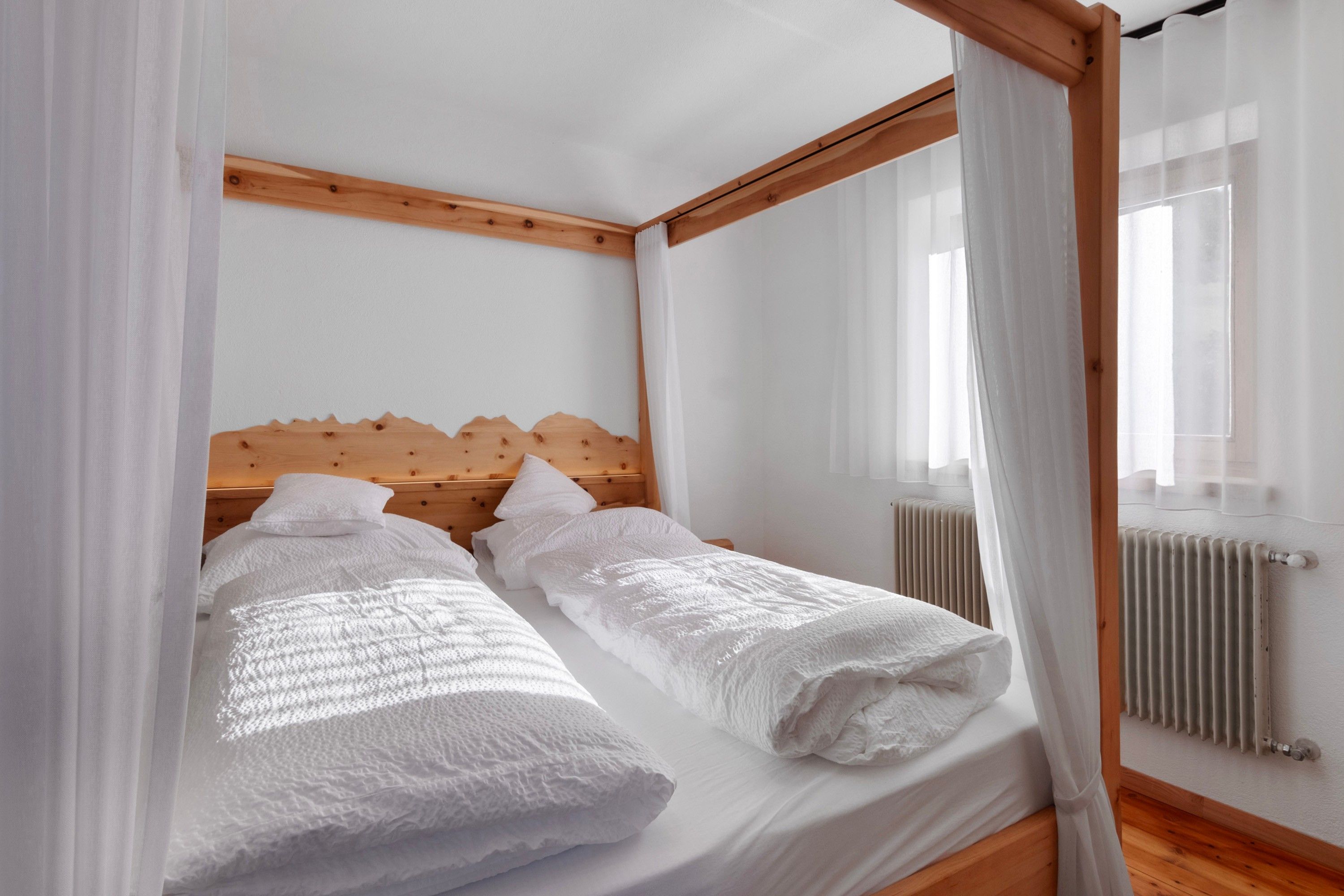 A double bed with a wooden headboard, white bedding, and sheer curtains.