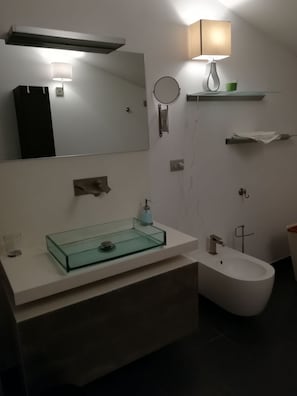 Un baño moderno con un lavamanos de vidrio, un inodoro, un bidé, un espejo y un toallero de pared.