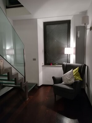 Un interior moderno con una escalera, un ventanal con persiana texturizada, un sillón con cojines y una lámpara de pie.
