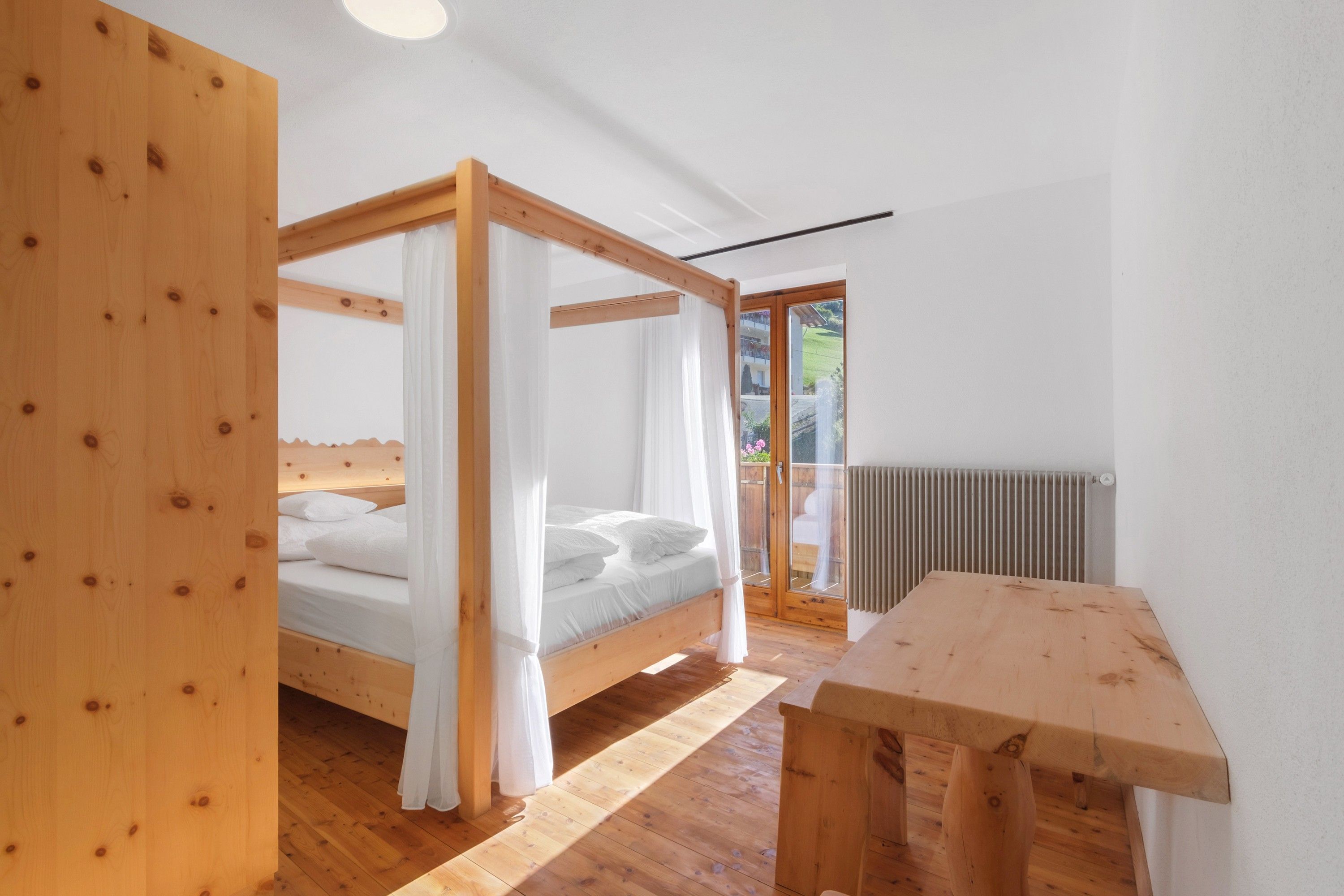 A wooden four-poster bed with white bedding, a wooden table, and a view of a garden through a glass door.