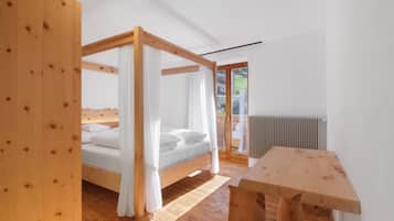 A wooden four-poster bed with white bedding, a wooden table, and a view of a garden through a glass door.