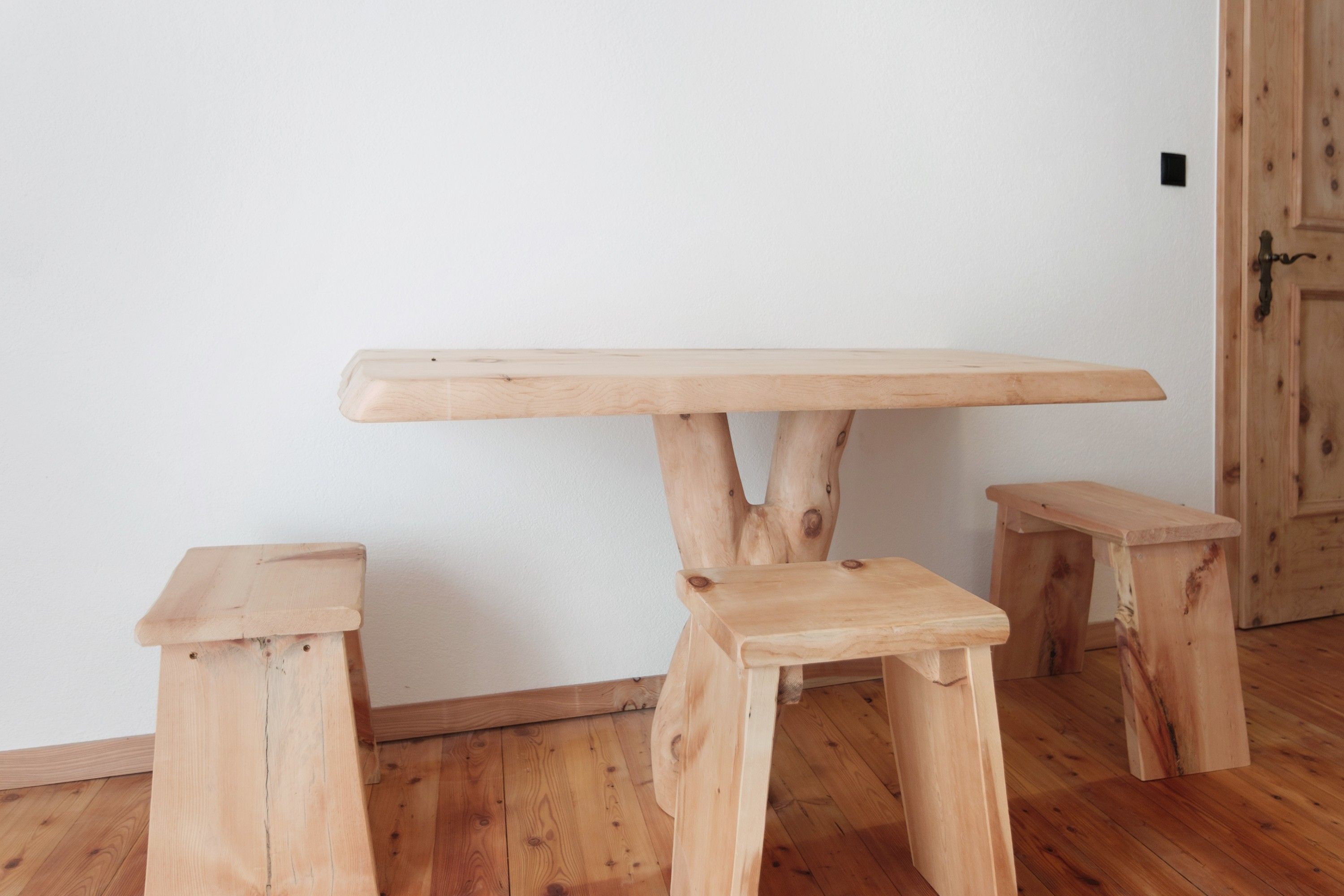 Ein schlichter Holztisch mit drei Beinen in einem Zimmer mit Holzboden und weißer Wand.