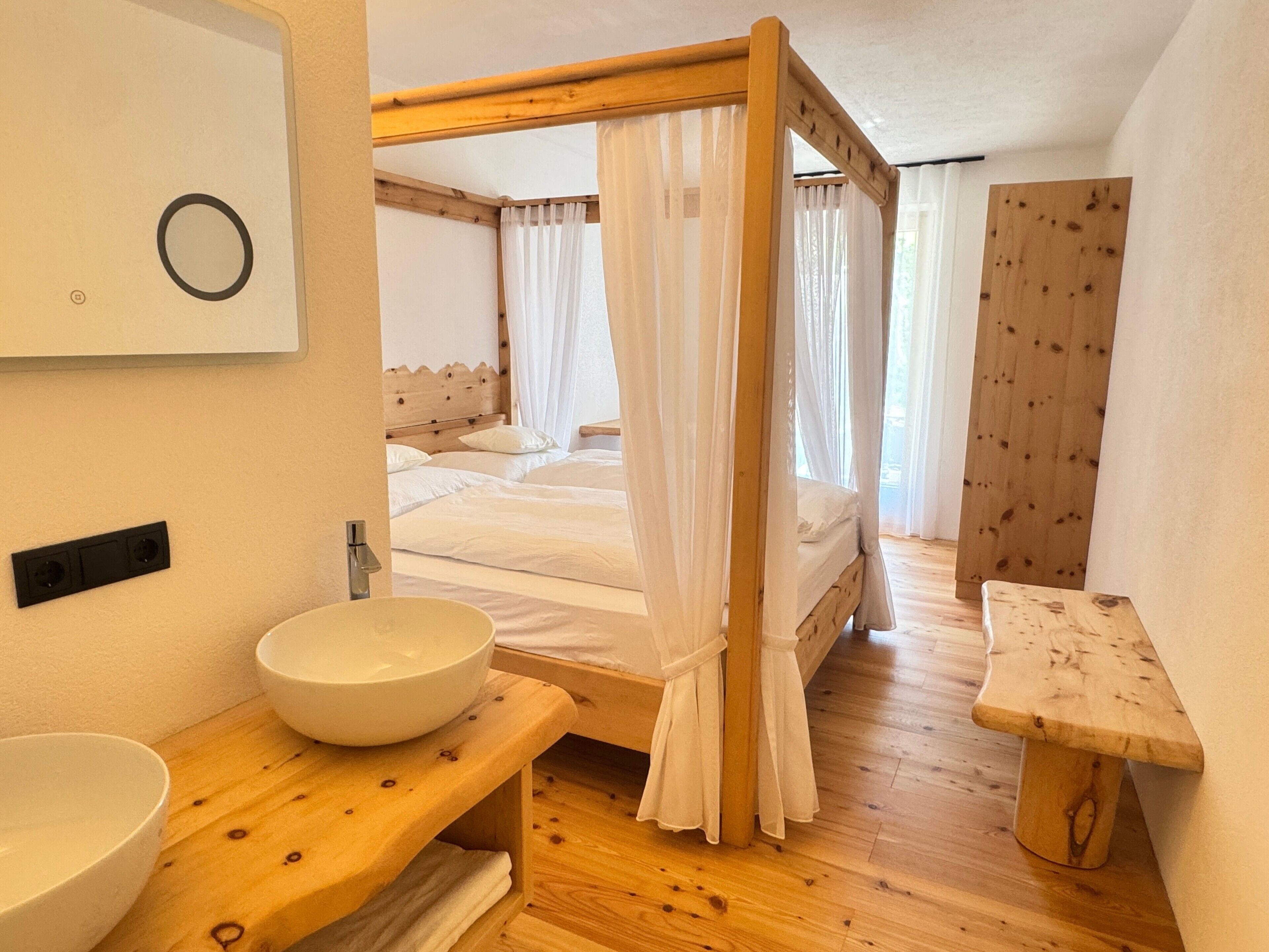 Ein Schlafzimmer mit Himmelbett, Nachttisch aus Holz, weißem Waschbecken und einer Holzbank.