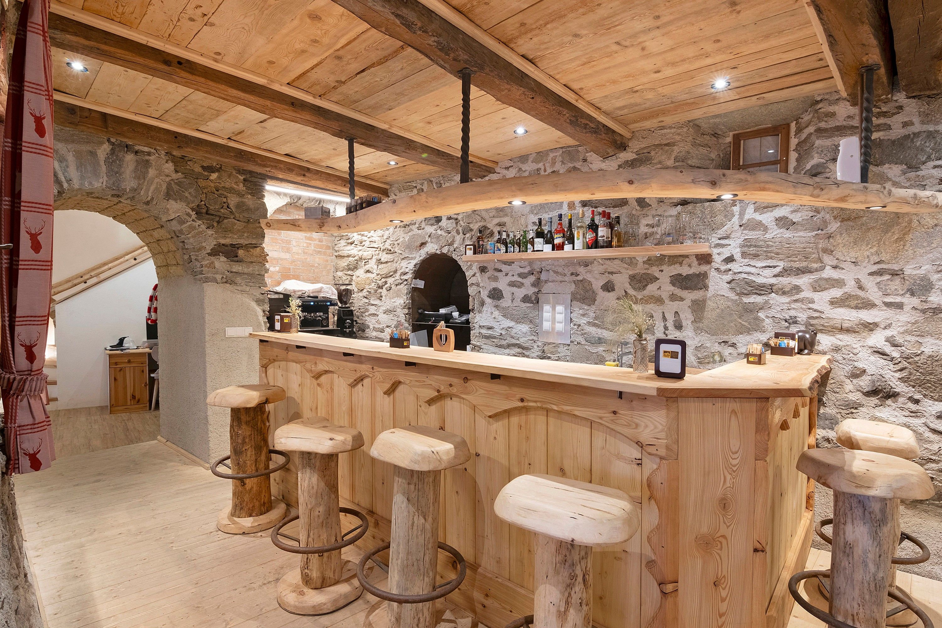 A rustic bar area with wooden stools, a stone wall backdrop, and a variety of bottles.