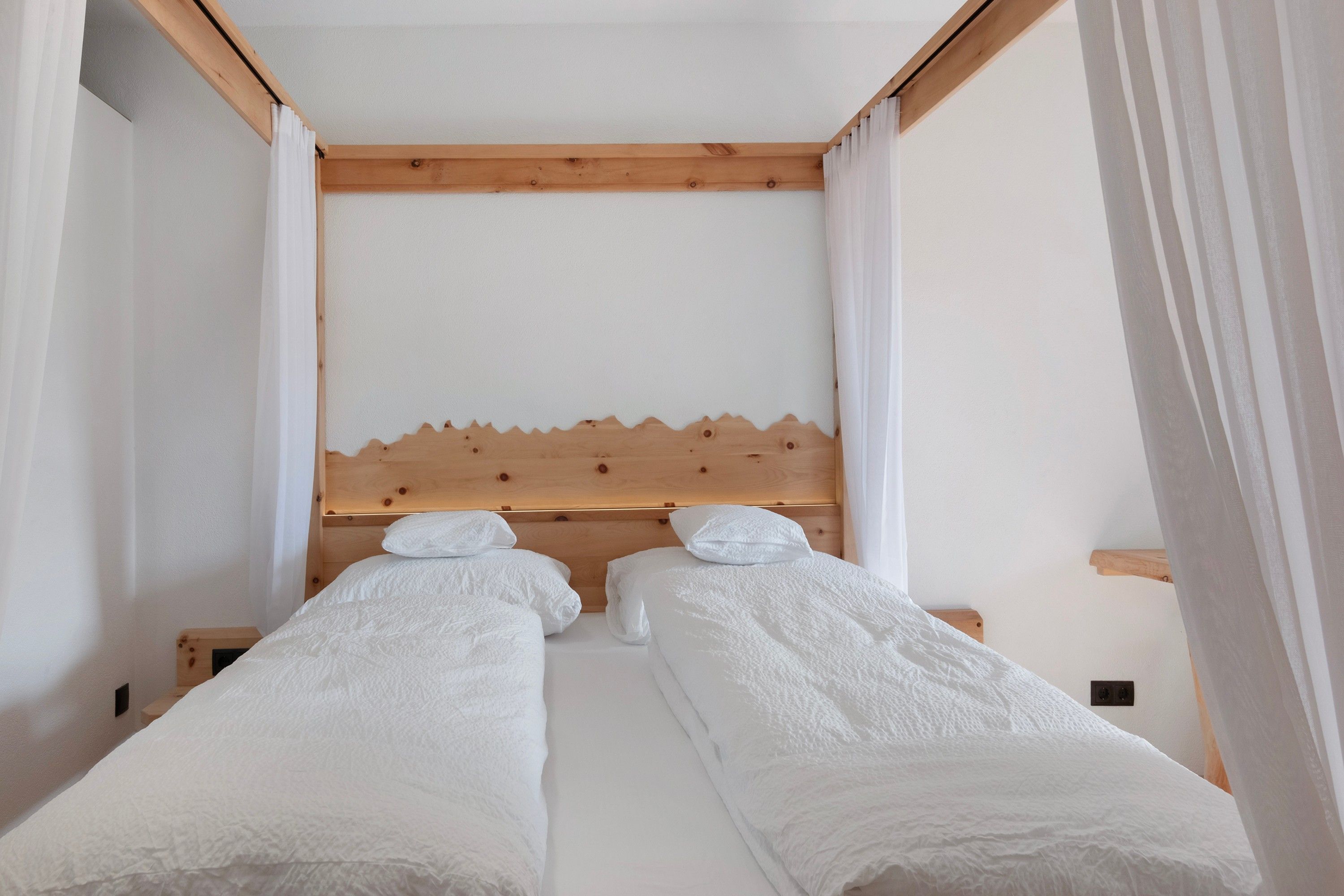 A room with two single beds, wooden headboards, white bedding, and sheer curtains.