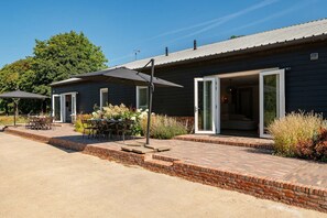 Ein Gebäude mit einer Terrasse, die einen Tisch und Stühle, eine überdachte Sitzgelegenheit und einen Ziegelweg zum Eingang bietet.