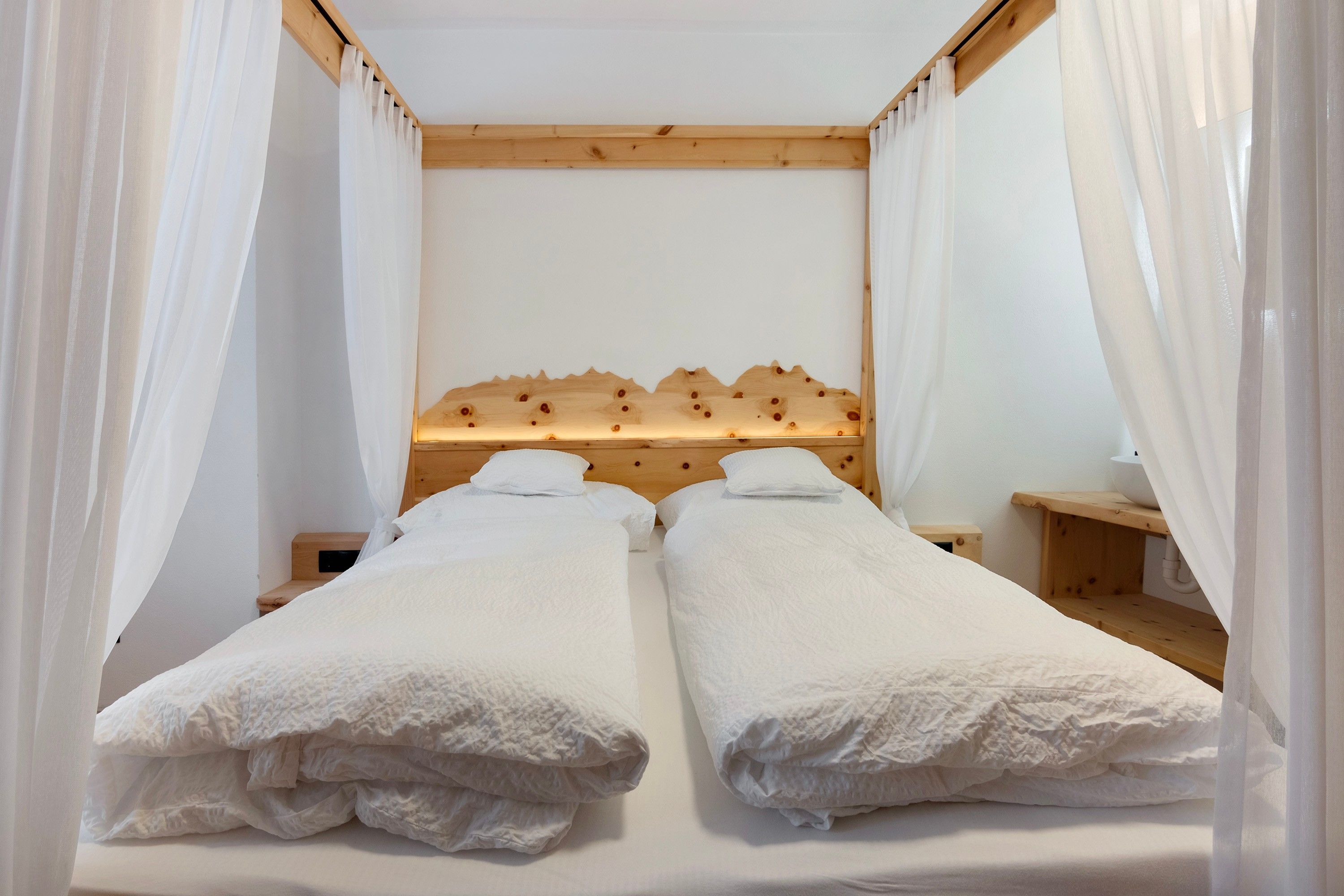 A double bed with a wooden headboard, white bedding, and sheer curtains.