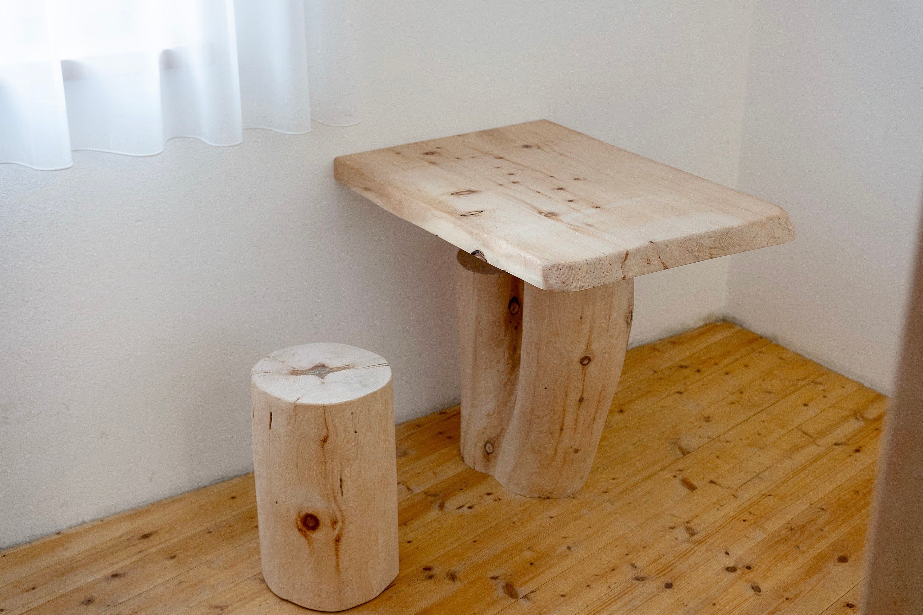 Ein kleiner Holztisch mit einem runden Hocker daneben, beide auf einem Holzboden vor einer weißen Wand.