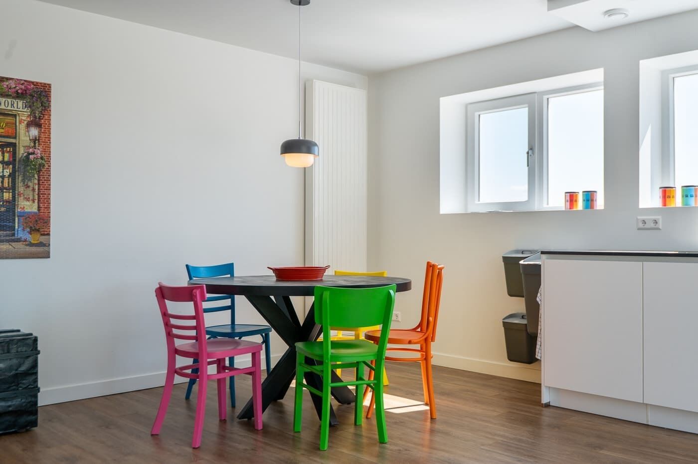 A dining area with a table and colorful chairs, a pendant light, and a kitchen area with cabinets and a window.