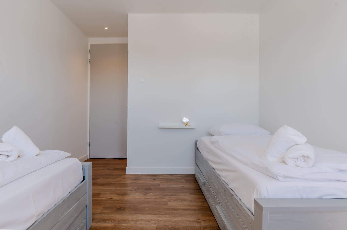 Ein Hotelzimmer mit zwei Betten, Holzboden, einer weißen Wand und einer kleinen Ablage mit einem dekorativen Gegenstand.