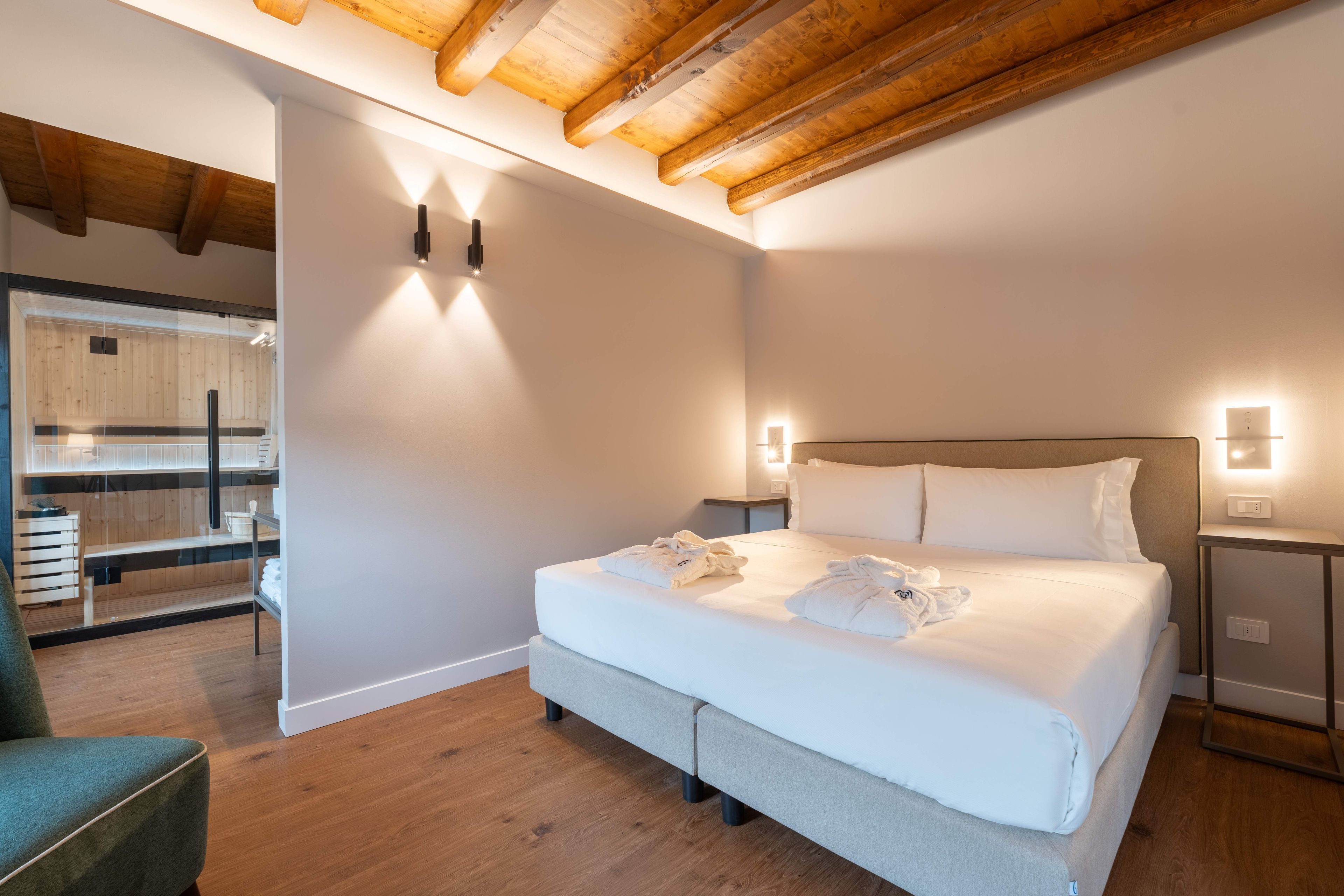 Moderni hotellihuone, jossa on suuri sänky, puupalkit katossa ja lasiseinä, joka jakaa huonetta.