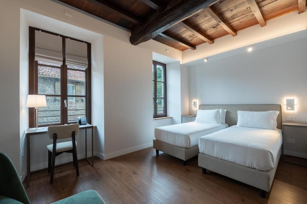A hotel room with two beds, a desk, a chair, and a window with a view of greenery.