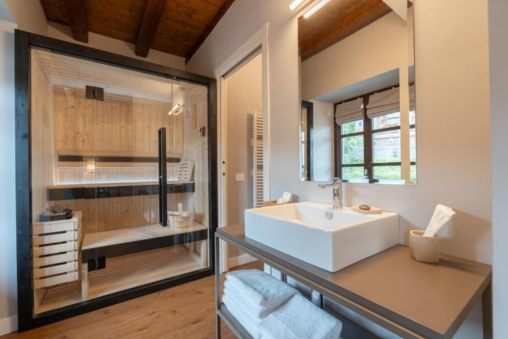 A modern bathroom with a large white sink, a glass shower enclosure, and a wooden bench.