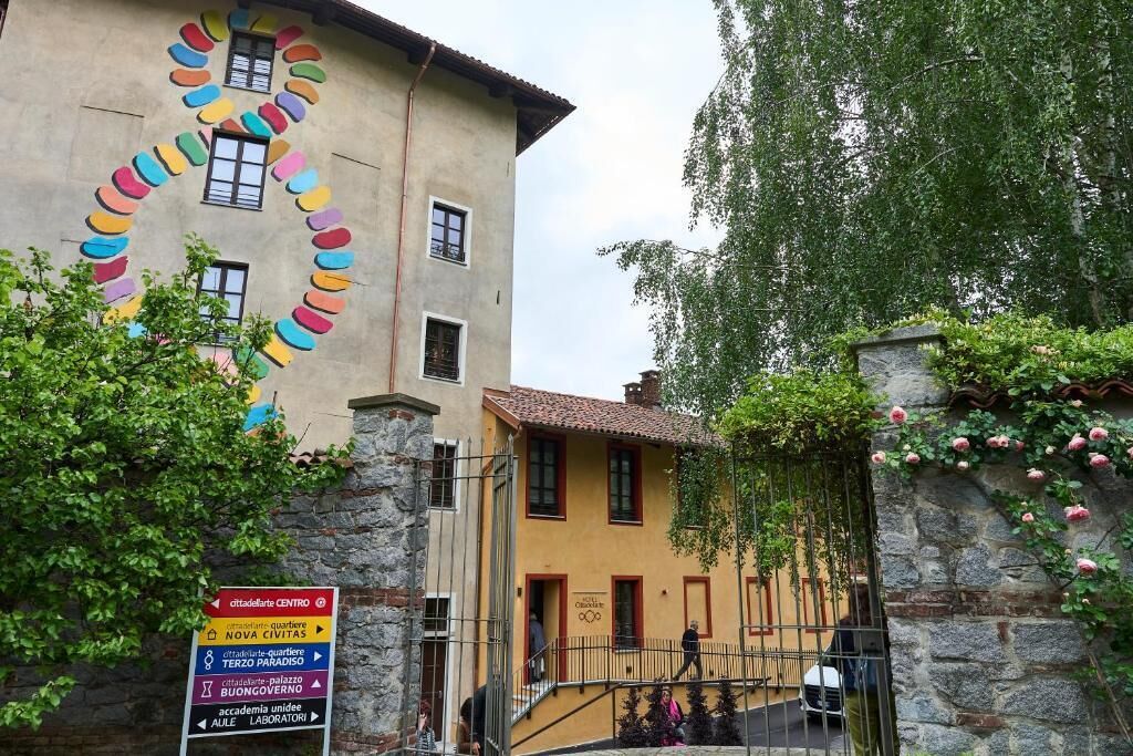 Un bâtiment dont la façade présente un motif circulaire aux couleurs vives, un mur de pierre orné de plantes grimpantes et un panneau indiquant les directions vers différents lieux.