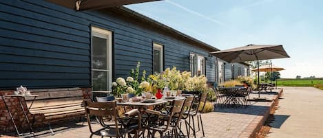 Een terras buiten met tafels en stoelen, een pad van stenen en een gebouw met grote ramen.