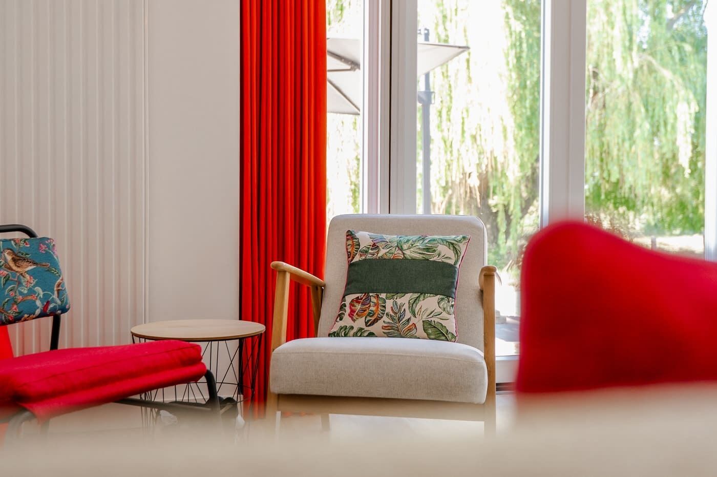 A room with a window, a chair with a floral pillow, a small round table, and a red chair.