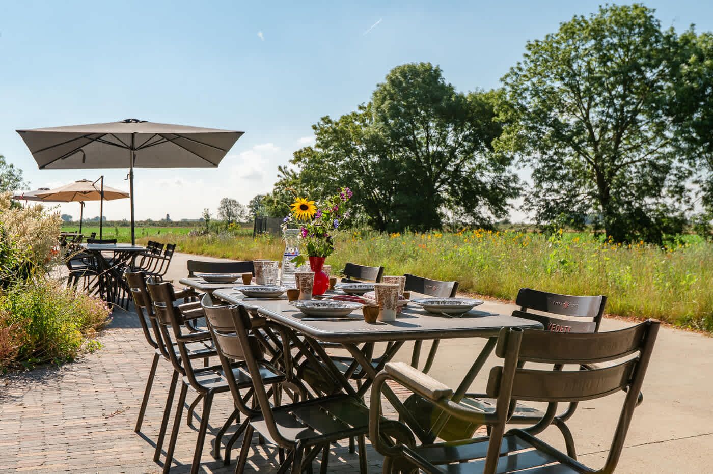 An outdoor dining area with tables and chairs, a large umbrella, and a vase with flowers.