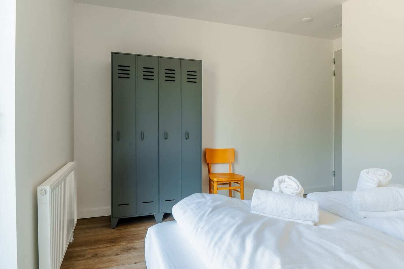 A hotel room with two beds, a wooden chair, and a set of grey lockers.