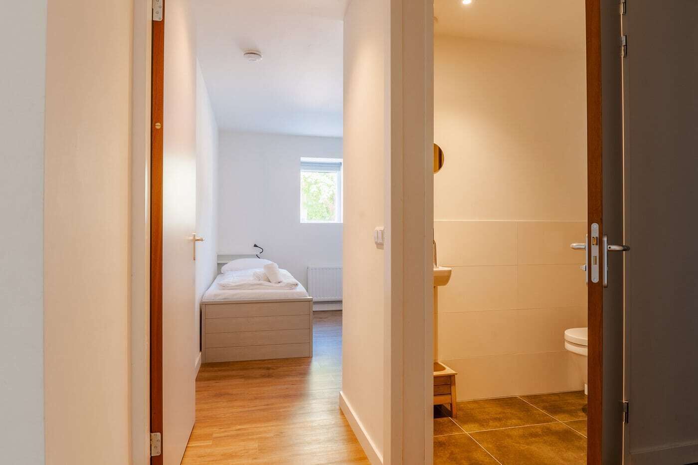 Une chambre d’hôtel avec un plancher en bois, un lit recouvert d’une couette blanche, une petite fenêtre et une salle de bain équipée de toilettes et d’un lavabo.
