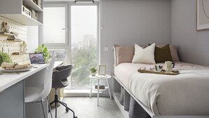 A modern bedroom with a bed, desk, and chair, a small side table, and a view of the city through a large window. - Student Only Bridle Works (Glasgow)