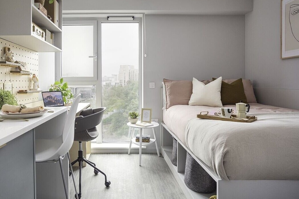 A modern bedroom with a bed, desk, and chair, a small side table, and a view of the city through a large window.