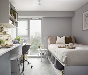 A modern bedroom with a bed, desk, and chair, a small side table, and a view of the city through a large window. - Student Only Bridle Works (Glasgow)
