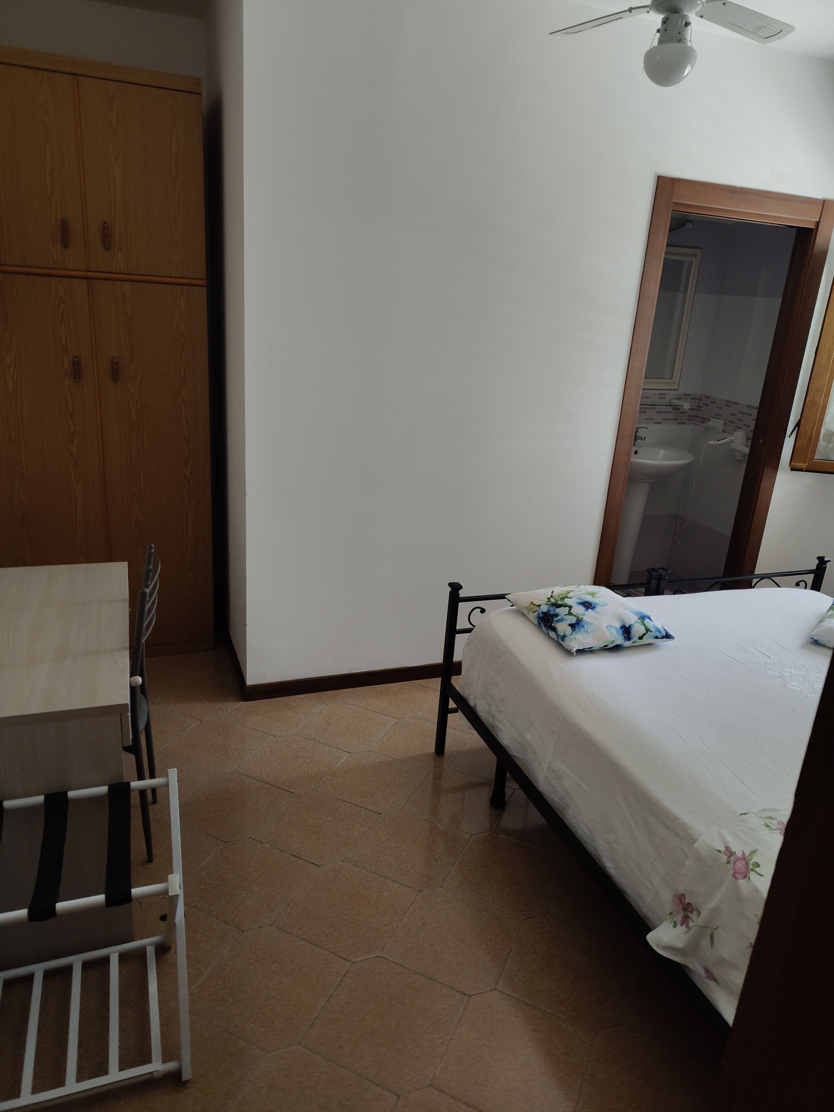 Une chambre à coucher comprenant un lit, une chaise, une armoire en bois, un ventilateur de plafond et une salle de bain visible à travers une porte ouverte.