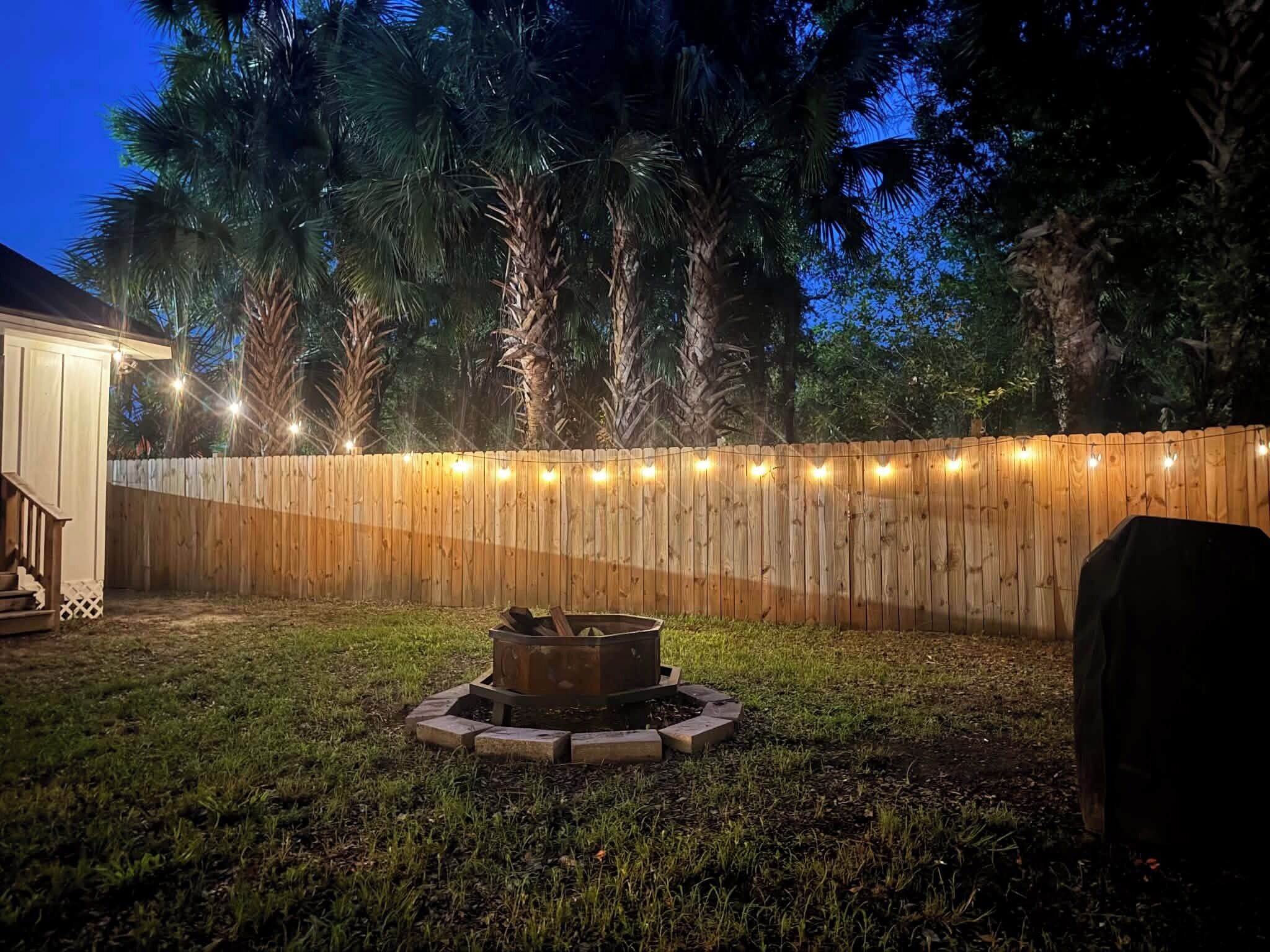 A backyard with a wooden fence, string lights, and a fire pit.