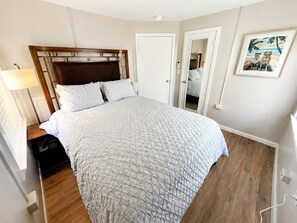 A bedroom with a wooden headboard, a bed with a quilted cover, a bedside table with a lamp, and a door leading to another room. - Cozy 2-bedroom house in beautiful Bay St. Louis with AC (Bay St. Louis)