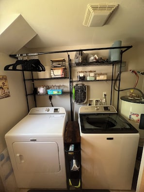 A laundry room with a front-loading washer and dryer, a metal shelving unit above, and a vent on the ceiling. - Cozy 2-bedroom house in beautiful Bay St. Louis with AC (Bay St. Louis)