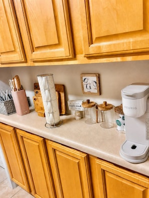A kitchen counter with wooden cabinets, a coffee maker, and various kitchen utensils and containers. - Cozy 2-bedroom house in beautiful Bay St. Louis with AC (Bay St. Louis)