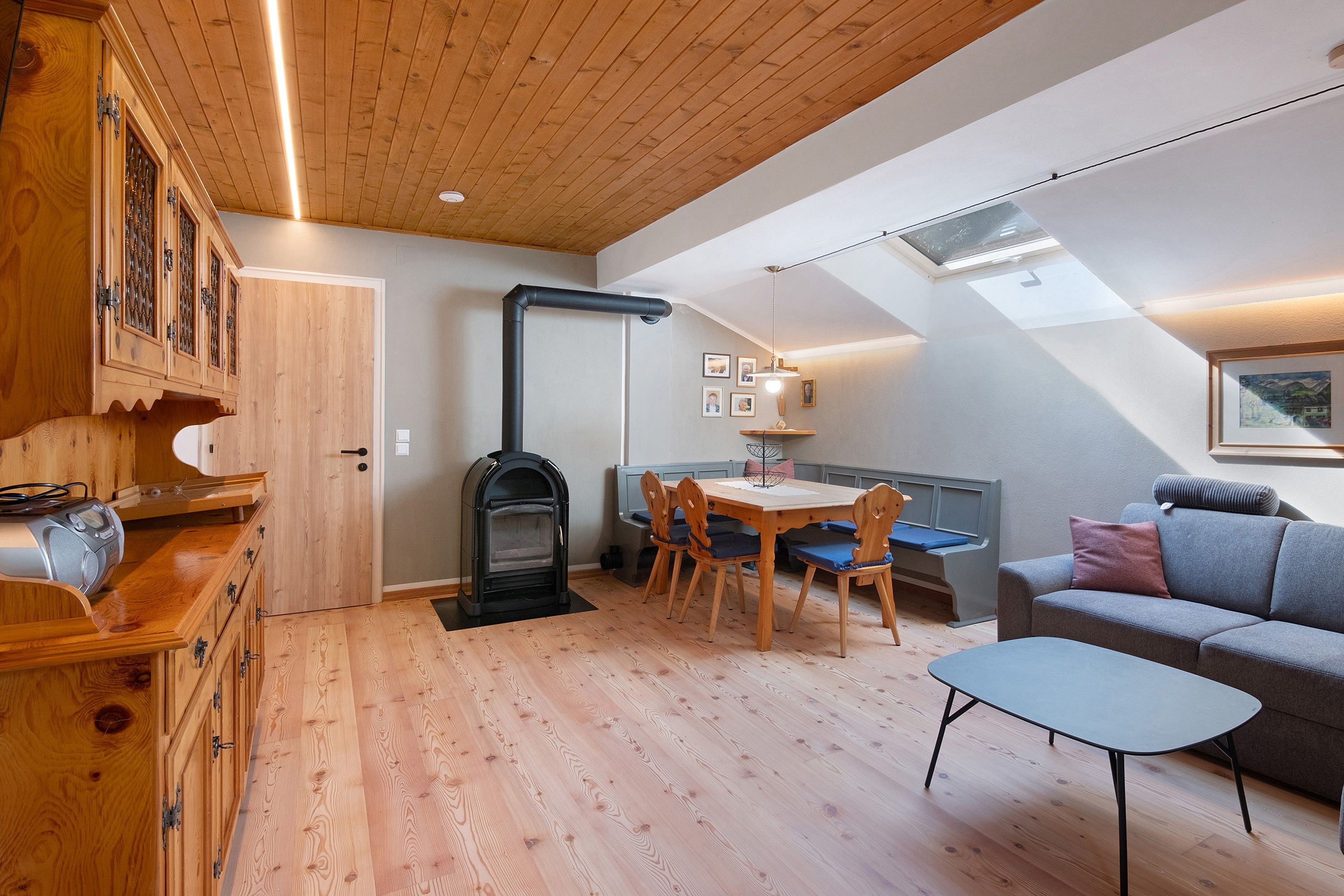 A cozy room with a wooden ceiling, a built-in stove, a dining table with chairs, a sofa, and a small table.