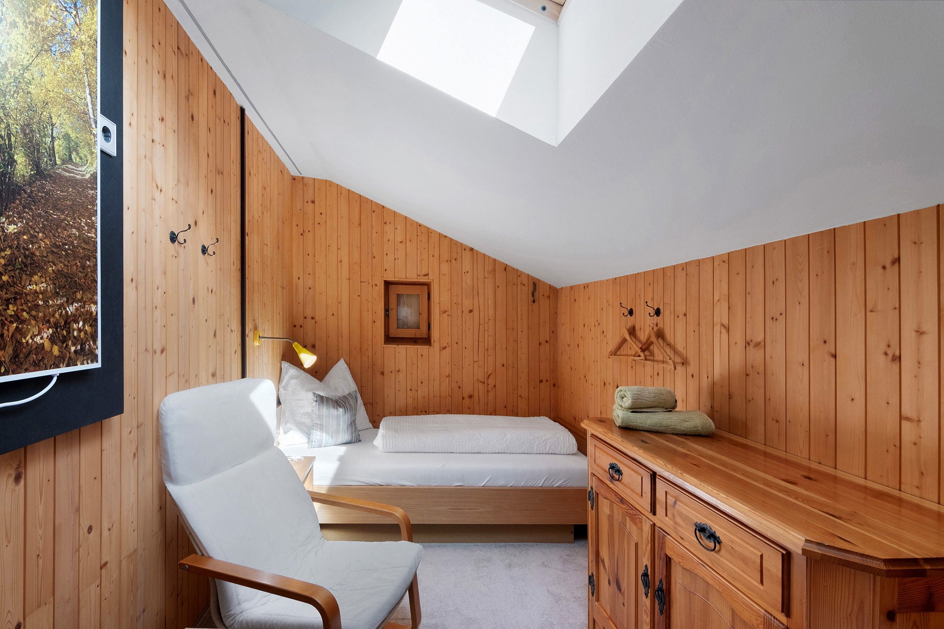 A bedroom with a sloped ceiling, a bed, a chair, a bedside table, and a window with a view of trees.