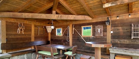 Una cabaña rústica de madera con comedor que incluye una mesa, bancos, una chimenea de piedra y vistas por una ventana.