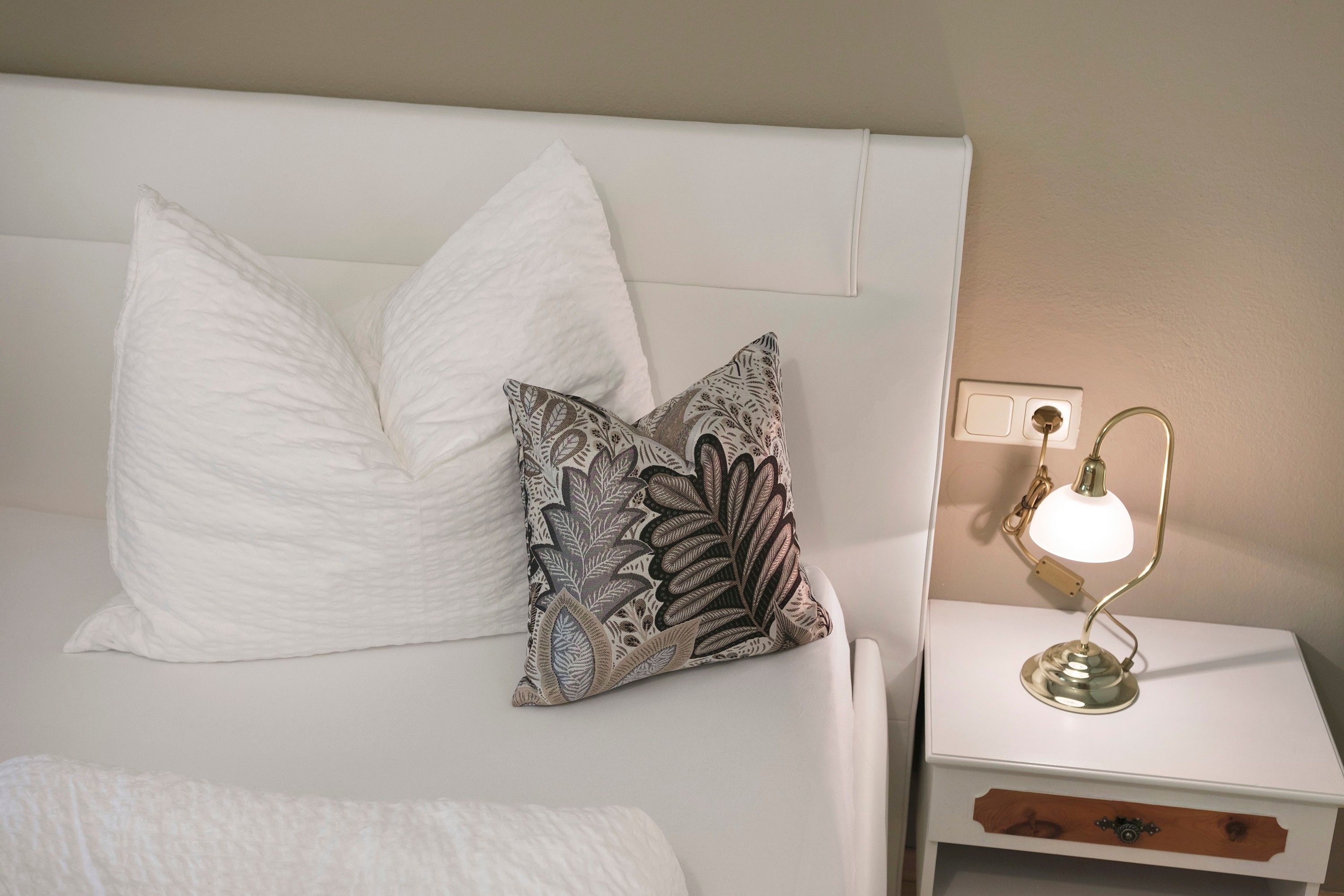 A bed with white pillows, a patterned pillow, and a bedside table with a lamp.